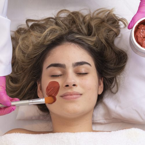 woman is lying in the cosmetologist s office on the procedure of moisturizing her face. The cosmetologist applies a moisturizing mask to the patient's face.