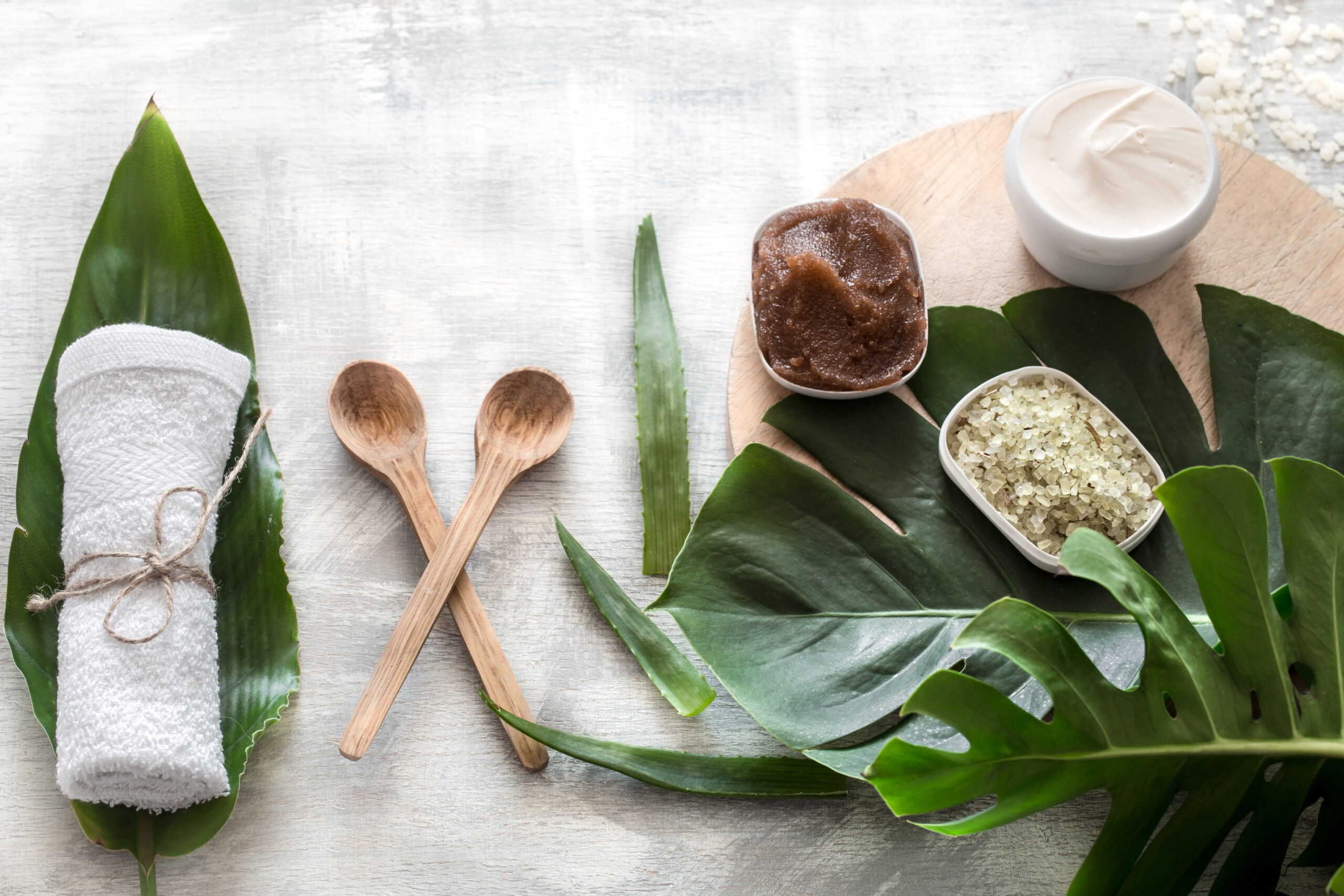 Flat-lay natural cosmetics and accessories with tropical leaves to cleanse the skin. Basic hygiene and body care items on white background, copy space.