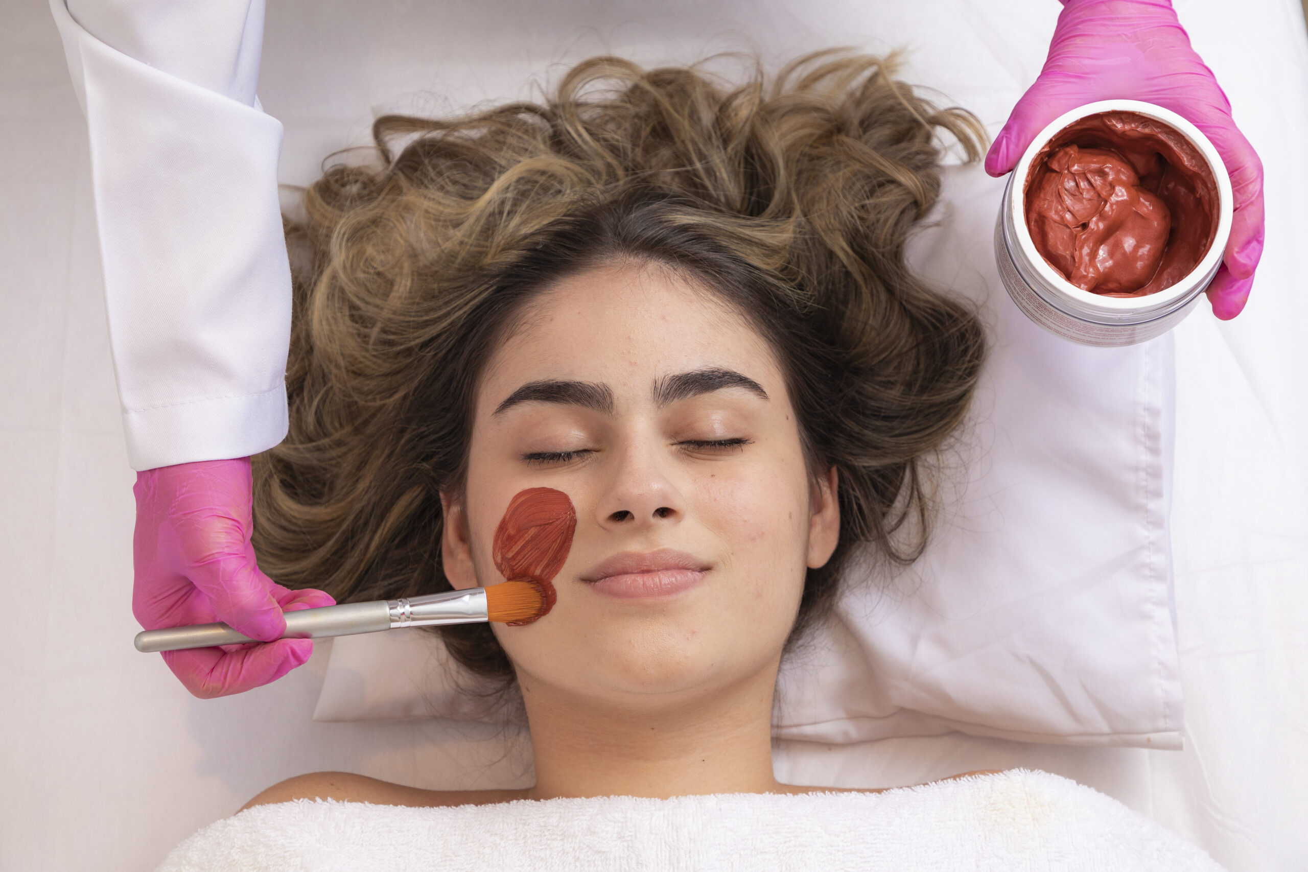 woman is lying in the cosmetologist s office on the procedure of moisturizing her face. The cosmetologist applies a moisturizing mask to the patient's face.